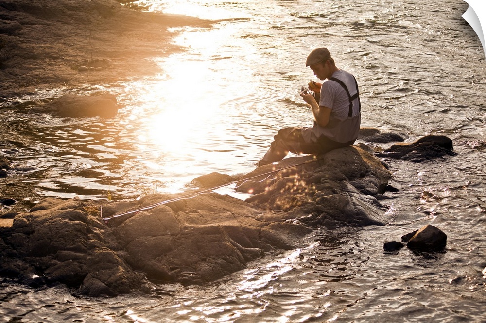 A young fisherman working on his rod and reel next to a river at sunset.