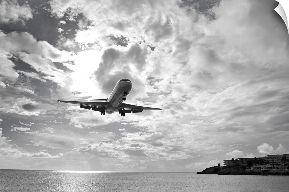 Black and white photograph taken of a commercial plane descending over ocean water about to land on an island.