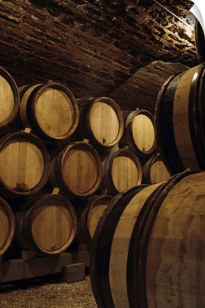 Vertical, oversized photograph of wooden barrels of wine stacked against the wall of a lit wine cellar.