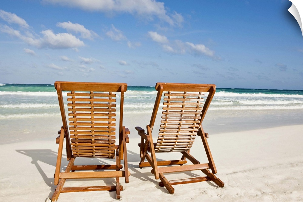 Large landscape photograph of two wooden lounge chairs sitting on the beach, beneath a blue sky.