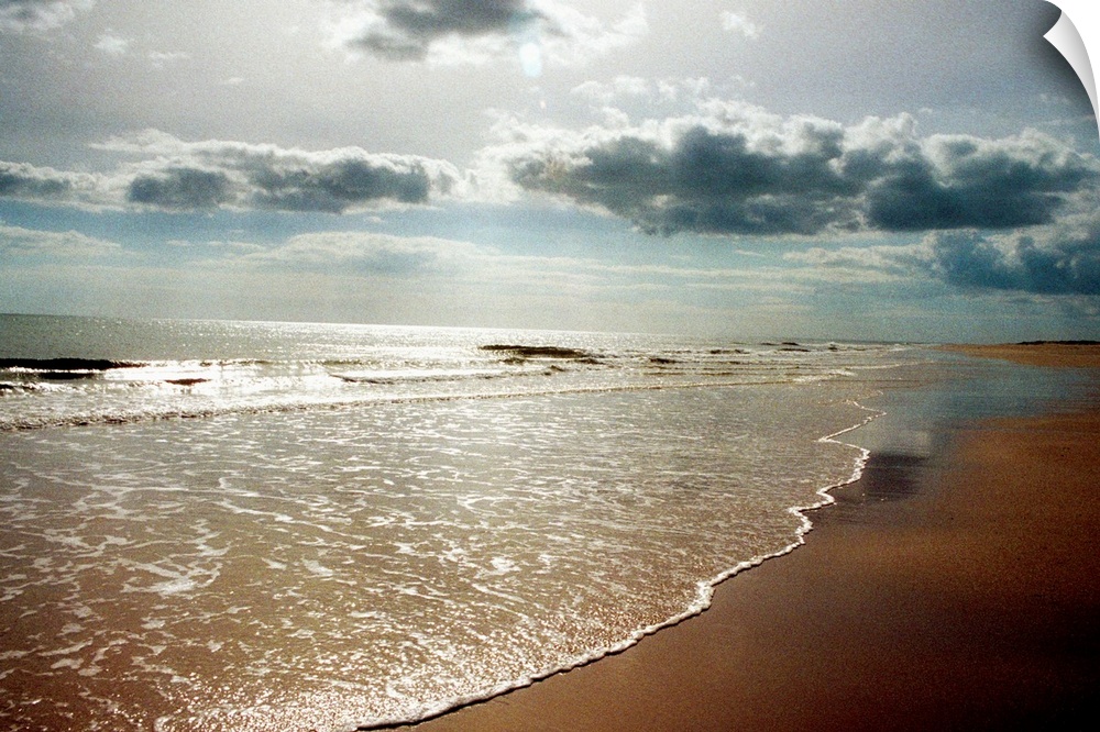 Sunlight shining down through puffy clouds onto a sandy beach and shallow ocean waves flowing onto the shore.