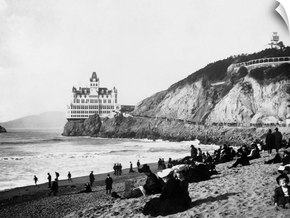 A crowd of people on the beach below the Cliff House enjoy the sun, the surf, and a view of Seal Rocks. The Gothic Revival...