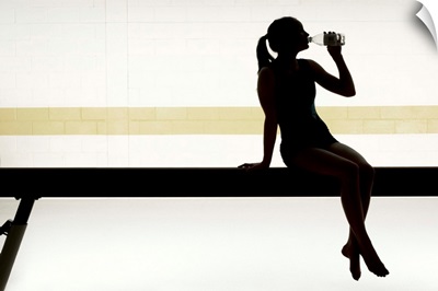 Female gymnast sitting on balance beam drinking water