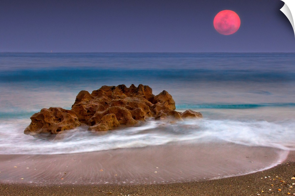A unique rock formation sits in the ocean water which is photographed under a deep red moon.