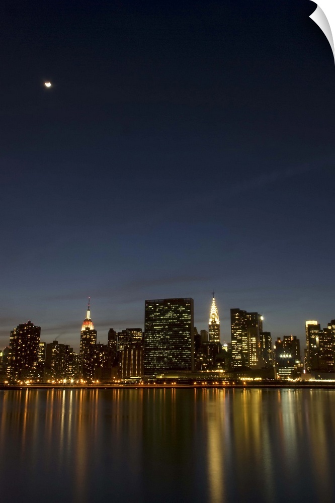 Midtown Manhattan at dusk with the moon above and reflection of the building lights in the East River.