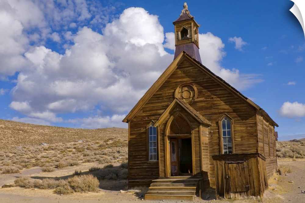 Large, horizontal photograph of a small, old wooden church surrounded by empty dessert, beneath a blue sky with fluffy clo...