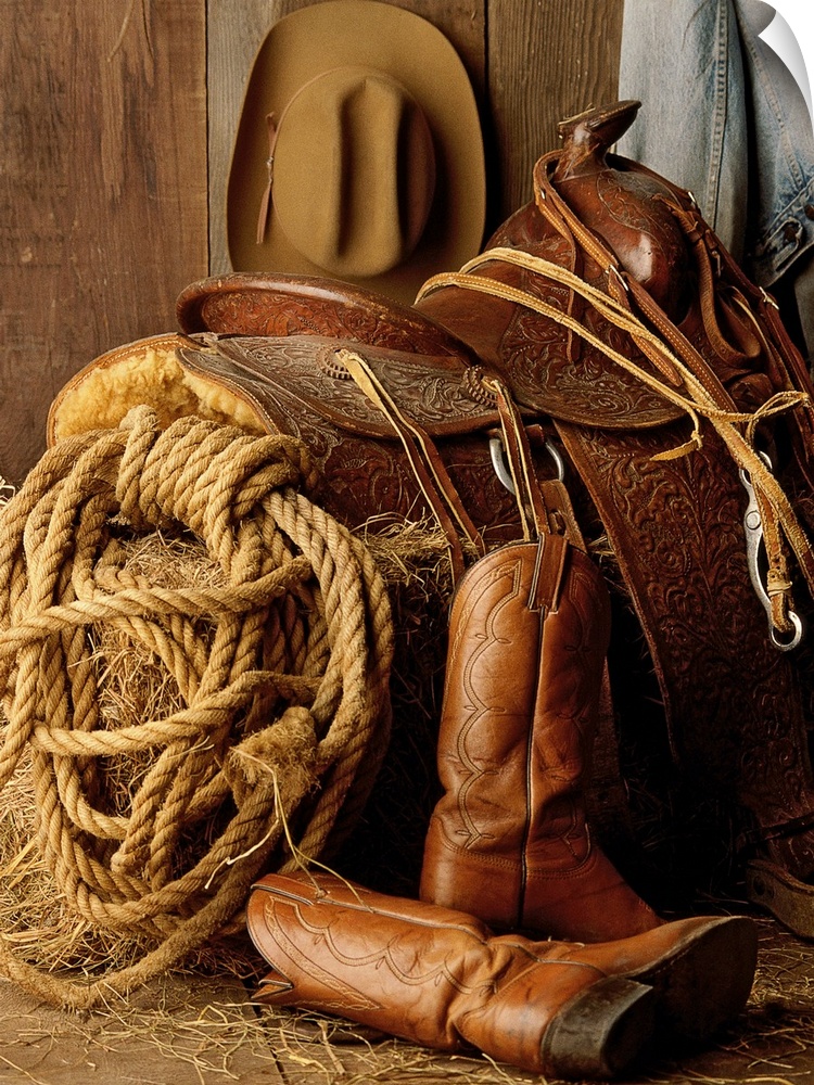 Staged photo of a horse saddle on a bale of hay surrounded by other Cowboy necessities such as a lasso, cowboy hat, and bo...