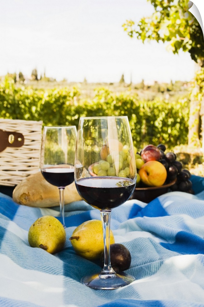Two glasses rest on a blanket, with pears, plums, grapes, and a loaf of bread with a sunny vineyard in the background.