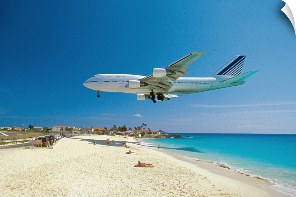 This is a landscape photograph of a passenger jet flying low over a tropical beach preparing to land.