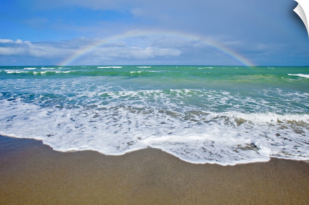 A large rainbow is photographed over the teal colored ocean.