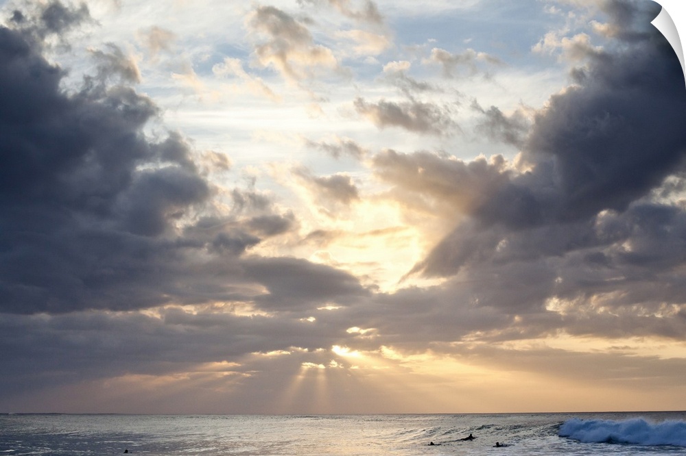 Crepuscular rays of light break through the sky over the blue ocean, surfers in the background