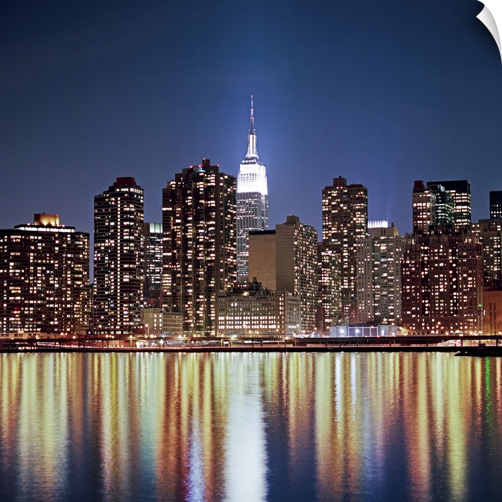 Photograph of "The Big Apple's" skyline and waterfront at dusk.  The buildings are lit up and lights are reflecting off th...