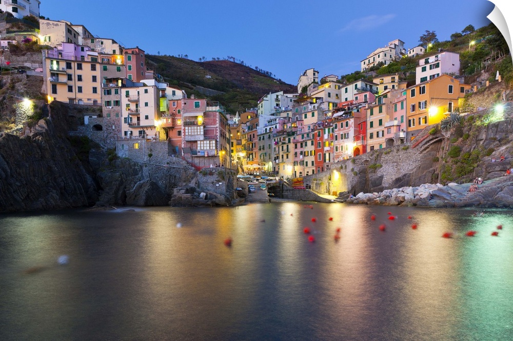 A town sits on a cliff and has an inlet of water in between. The buildings are lit up and lights reflect below.