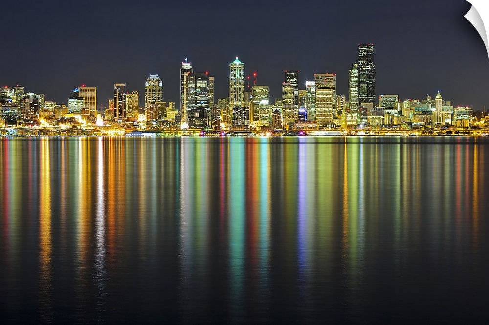 Large photograph taken of the Seattle skyline at night with the buildings lit up and reflecting in the water below.