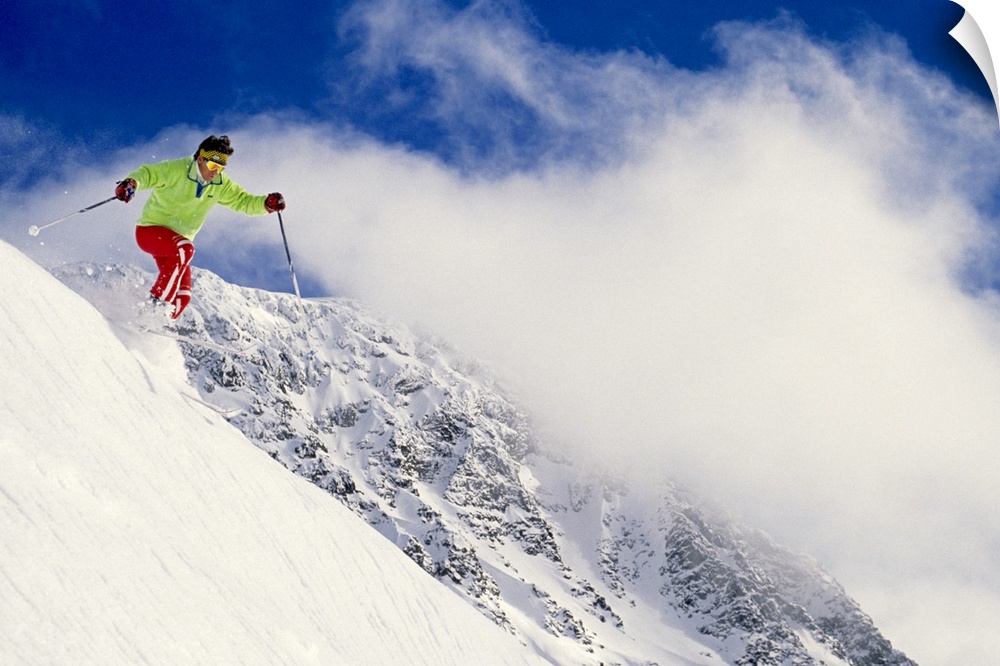 Photograph of man on skis coming down snowy slope with huge mountain in background under a cloudy sky.