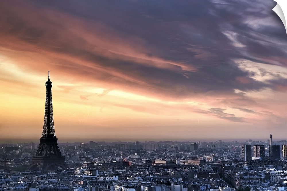 Big, horizontal photograph of the sun rising through wispy clouds as the Eiffel Tower looms over the city of Paris, France.