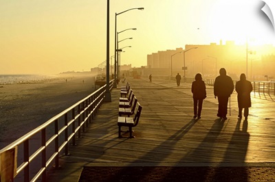 Three people walking down boardwalk near beach at sunset.