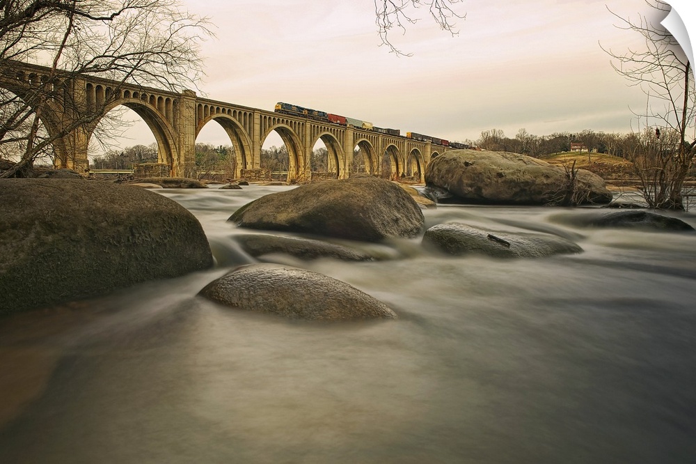 Large rocks sit on the banks for a river with a multi arched bridge spanning the river and a train running on top of the b...