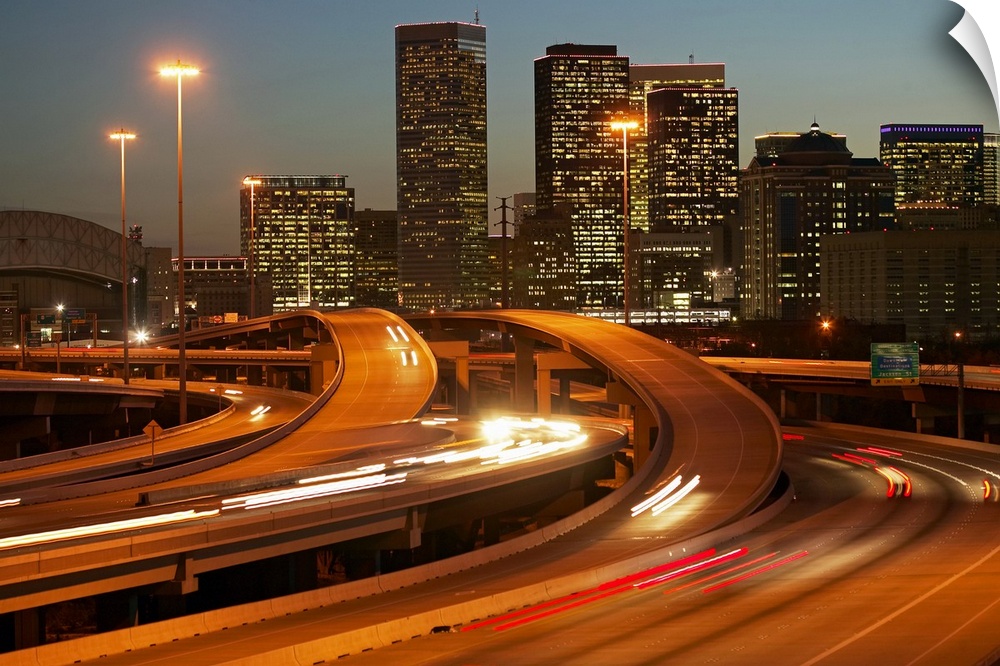 Photograph of the Houston skyline taken at night with the buildings lit up and cars lights streaking by on the highway.