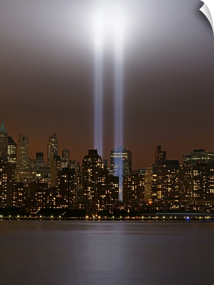 This vertical piece shows two beams of light shining up from where the twin towers stood. The skyline is lit up under a da...