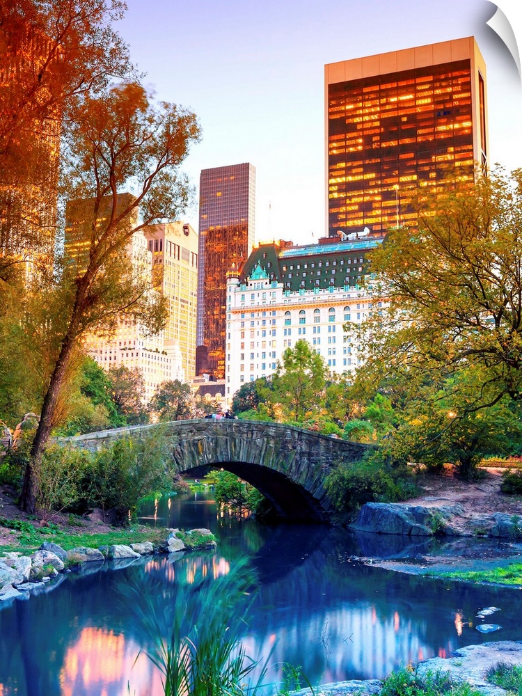 Vividly colored photograph of a bridge over a stream in Central Park, with skyscrapers in the distance.