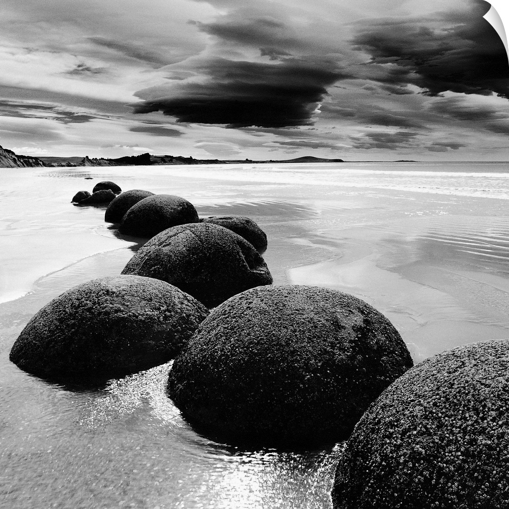 Black and white photograph of a row of smooth rocks on a beach with a dramatic sky full of clouds.