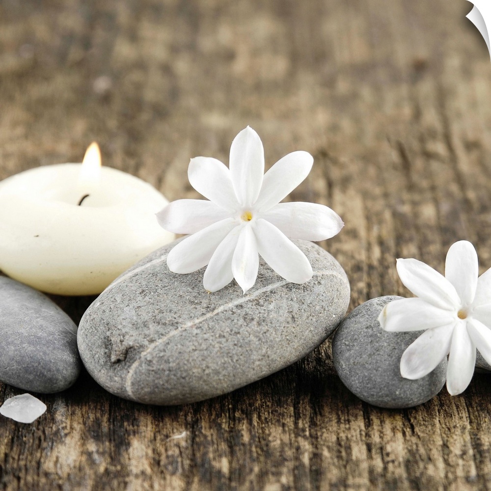 Square image of white flowers on smooth gray rocks with a candle on wood.