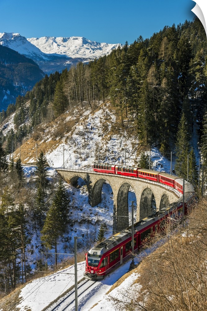 The famous red train of Albula mountain railway passing through a scenic winter alpine landscape near Filisur, Graubunden,...