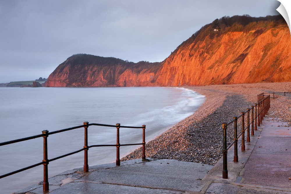 Early morning sunlight glows against the distinctive red cliffs of High Peak, viewed from the beach at Sidmouth, Devon, En...