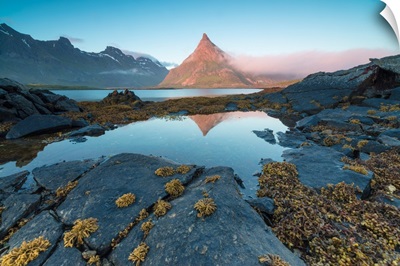 The rocky peak of Volanstinden lighted up by the midnight sun is reflected in blue sea