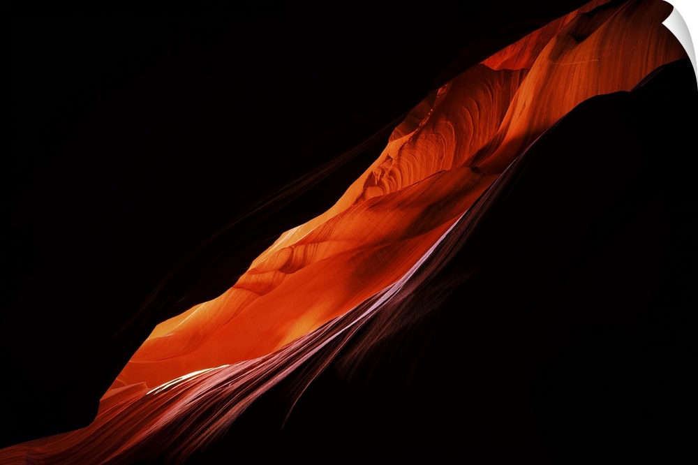 Large, landscape photograph of darkness surrounding a diagonal, thin area of a sandstone formation in Antelope Canyon, in ...