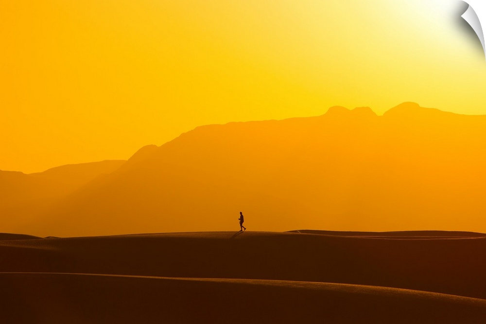 A lone man walks on top of sand dunes during sunset.