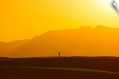 A lone man walks on top of sand dunes during sunset