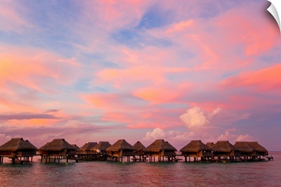 A vibrant pink and red sunset over bungalows on stilts over the water