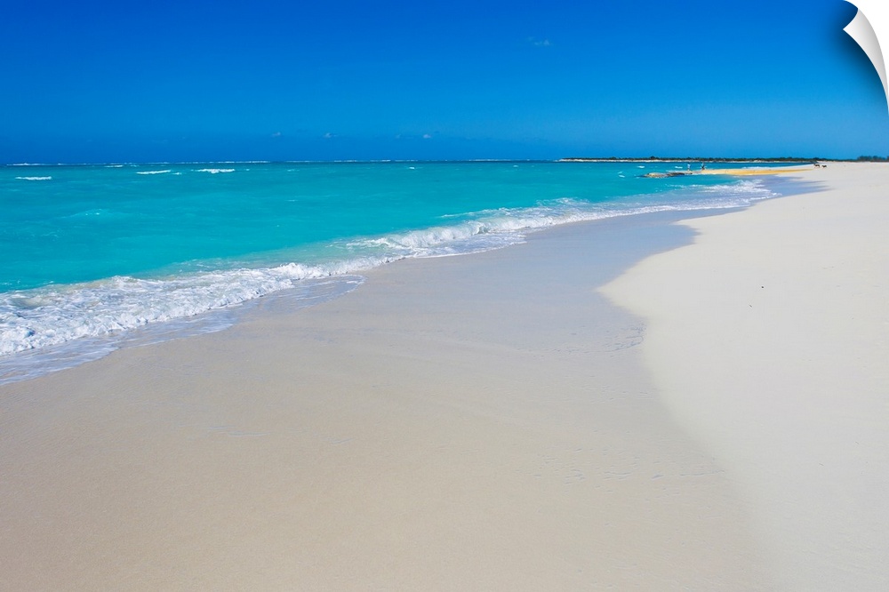 A beautiful picture of teal colored ocean water coming up onto the white sand beach. A clear blue sky hangs over.