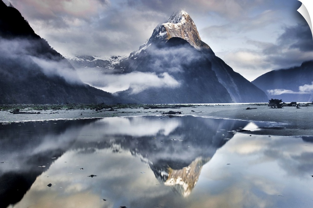 A landscape photograph of a snow covered mountain peak and misty clouds reflecting in shallow beach waters.