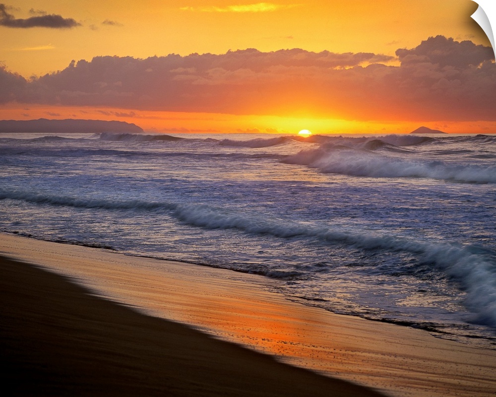 The sunset dips below the ocean horizon as ocean swells dash against the sandy tropical shoreline.