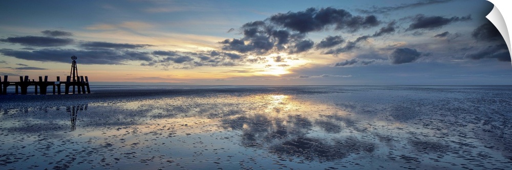 A panoramic image of a tranquil golden blue dawn with fluffy clouds over reflecting wet sand with a harbour installation.