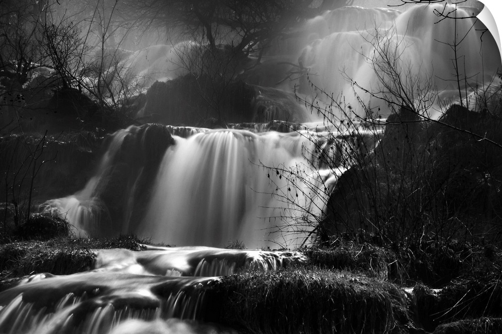 This black and white photograph is taken of several small waterfalls that are surrounded by bare trees and foliage.
