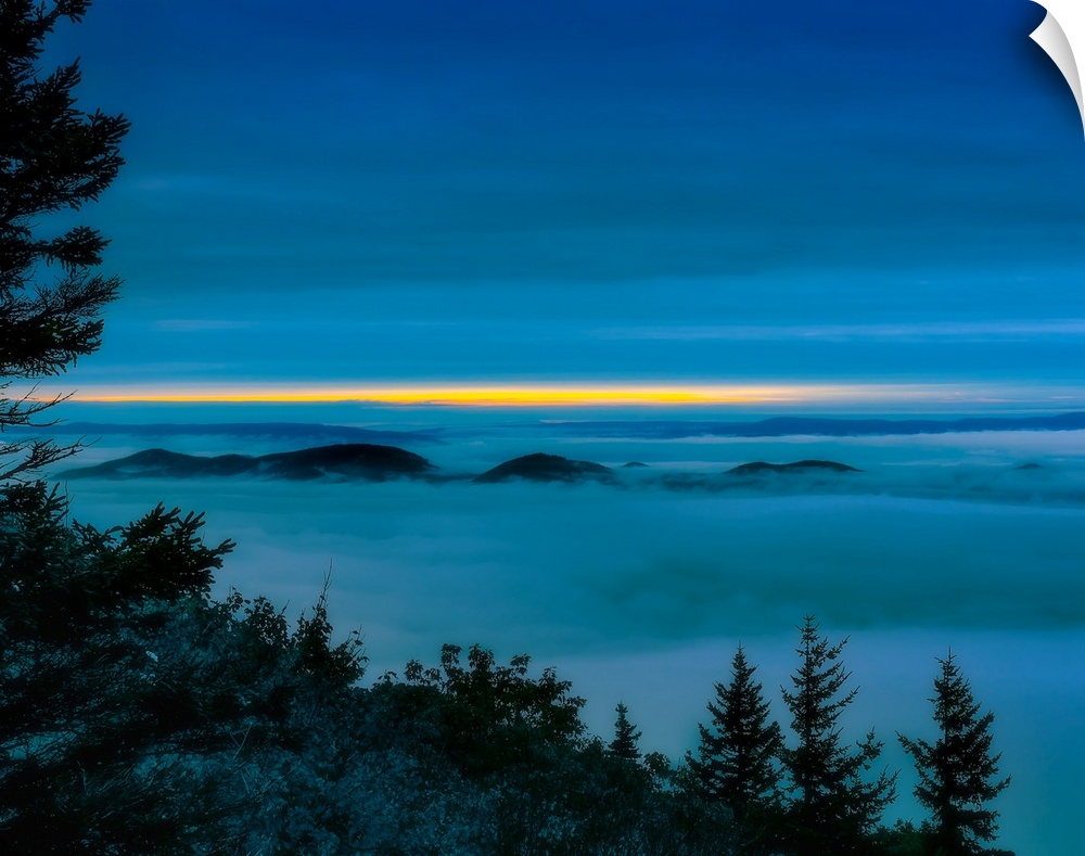 Cool toned photograph of the clouds covering the mountains with the sunrise peaking out in the distance.