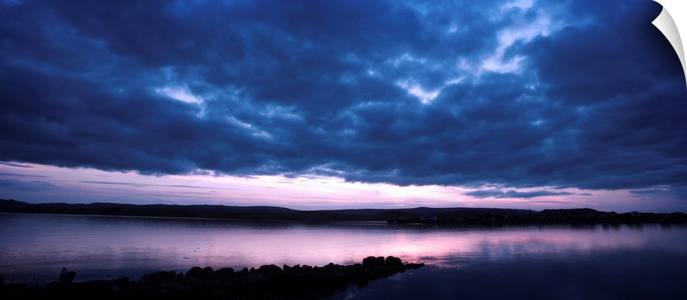 A dramatic panoramic skyscape over calm water in purples, blues and pinks with mountains beyond.