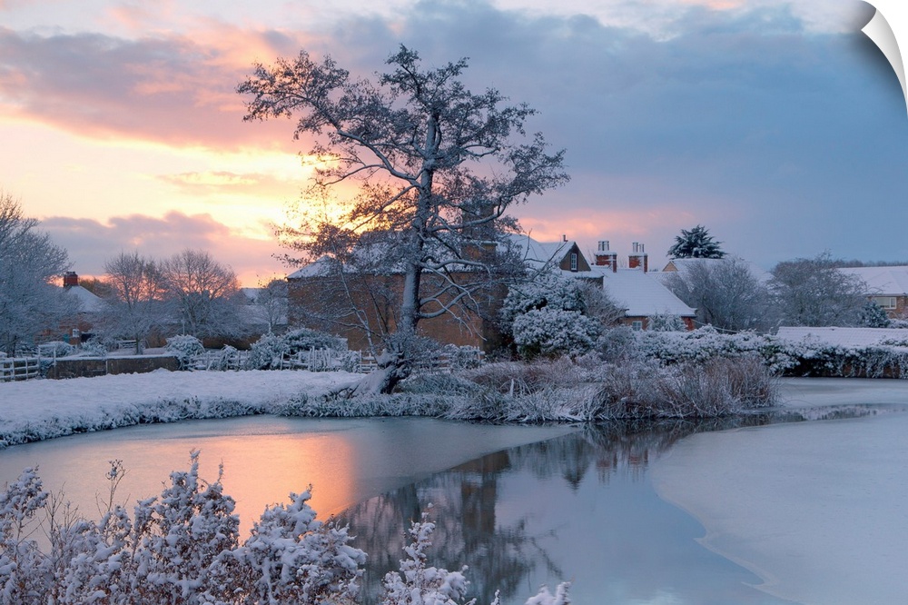 A golden dawn over a village pond and snowy fields to the village roofs beyond with trees in snow, Nottinghamshire, UK.