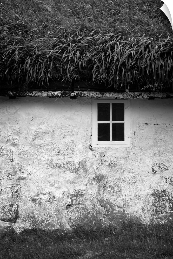 Black and white image of a small window in the stone wall of a house with a grassy roof.