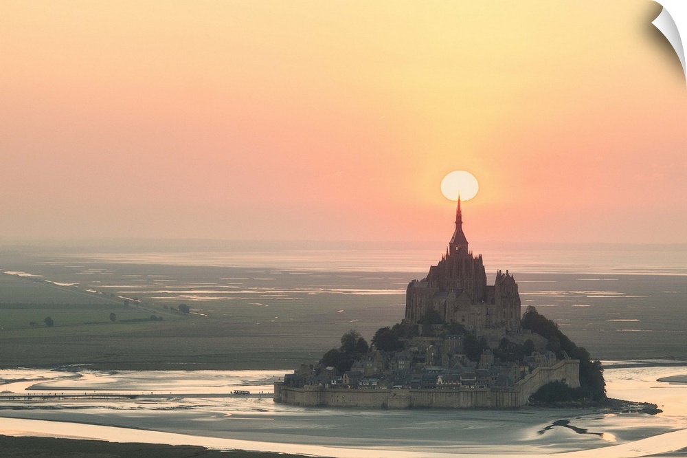 The sun appearing to rest on the spire at the top of Mont Saint Michel in France at sunset.