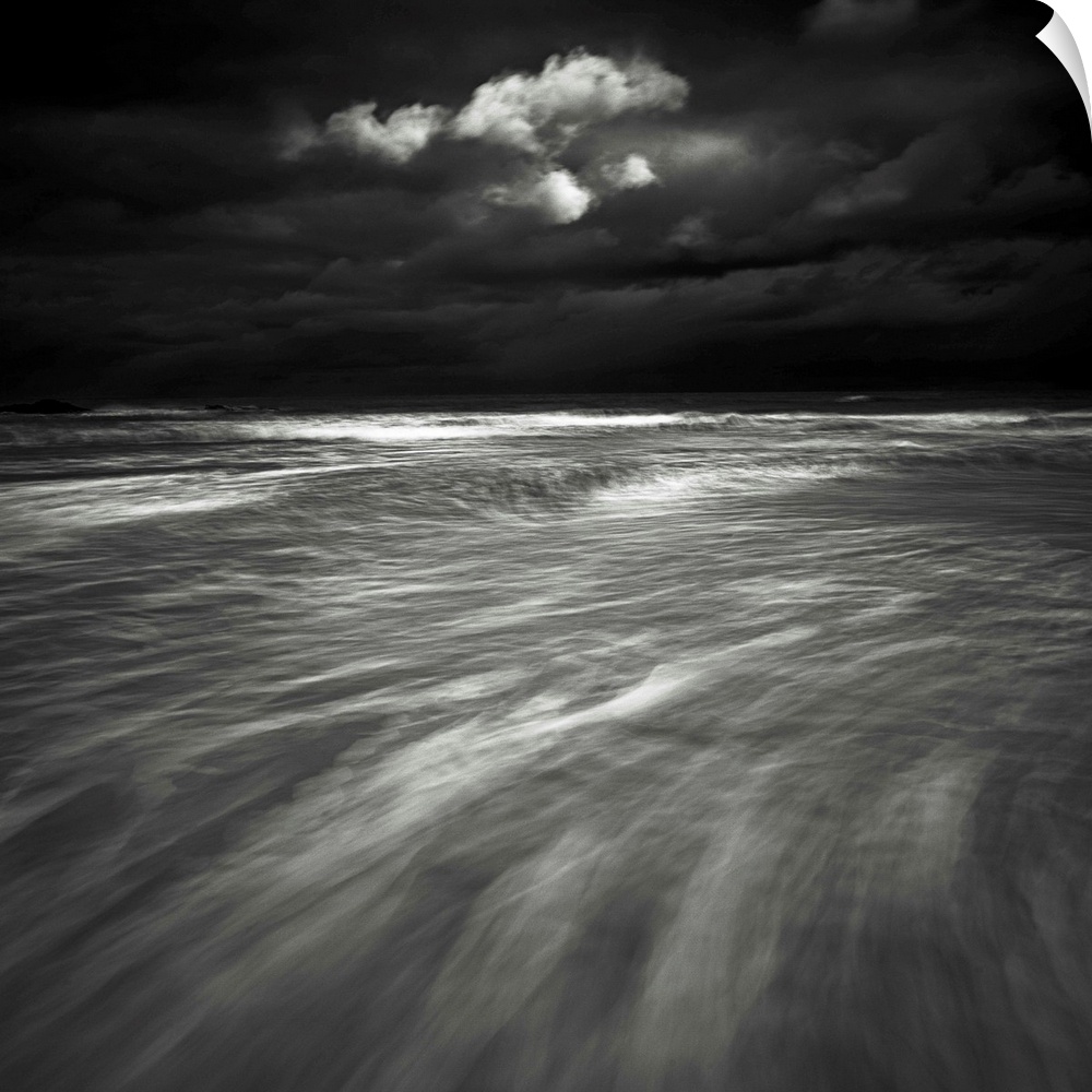 A monochrome black and white sepia toned dramatic image of swooshing wavws and a lone cloud in a dark stormy sky over a se...