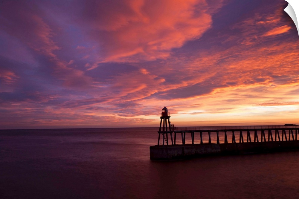 A dramatic peach, orange and red sunset over the lighthouse and pier at Whitby harbour, North Yorkshire, England.