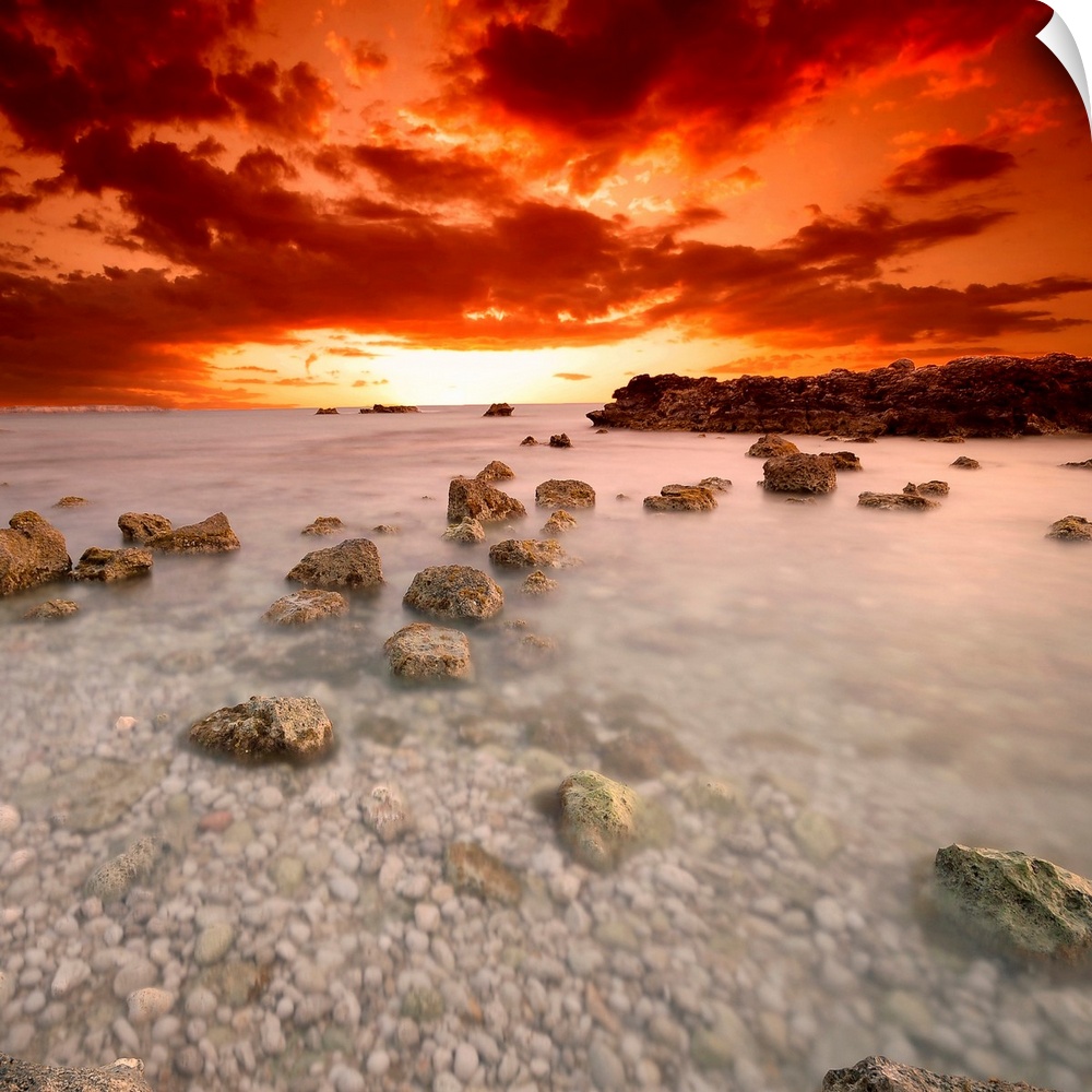 Square, fine art photograph of a shallow, large body of water full of rocks, beneath a deep orange and red sunset.