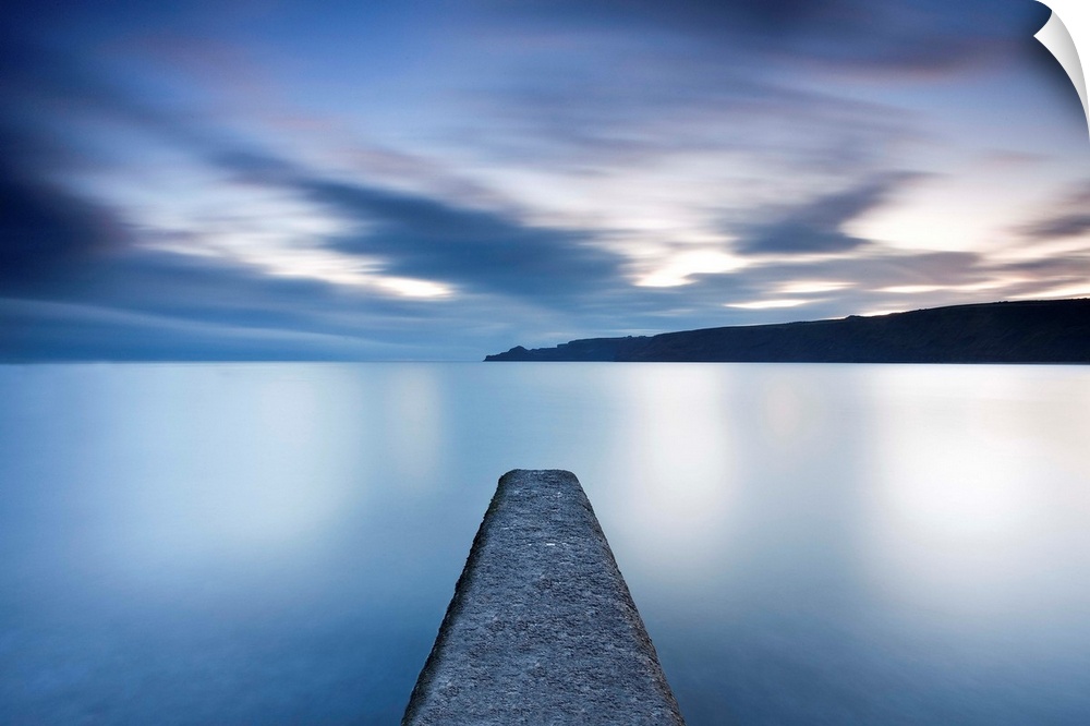 Tranquil calm blue seascape with symetrical jetty into a flat sea with floating clouds.