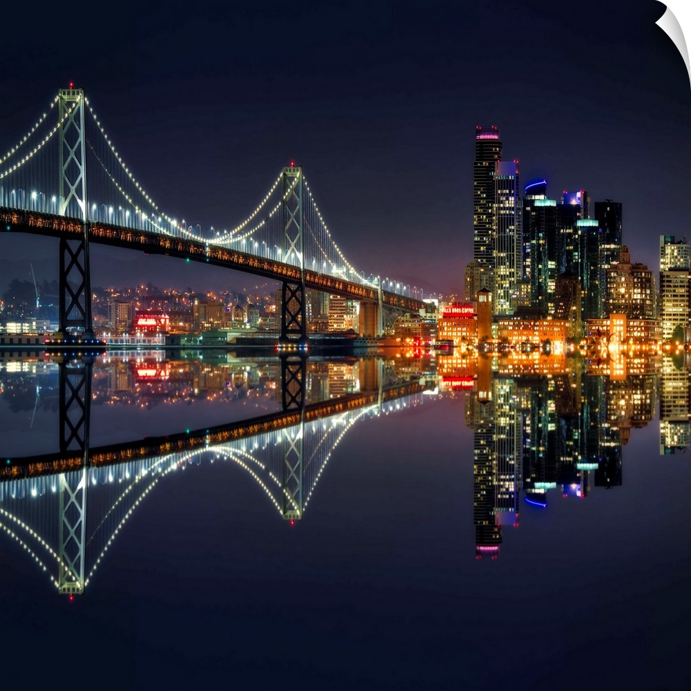 Square photograph of the Bay Bridge and downtown San Francisco lit up at night and reflecting onto the motionless water.