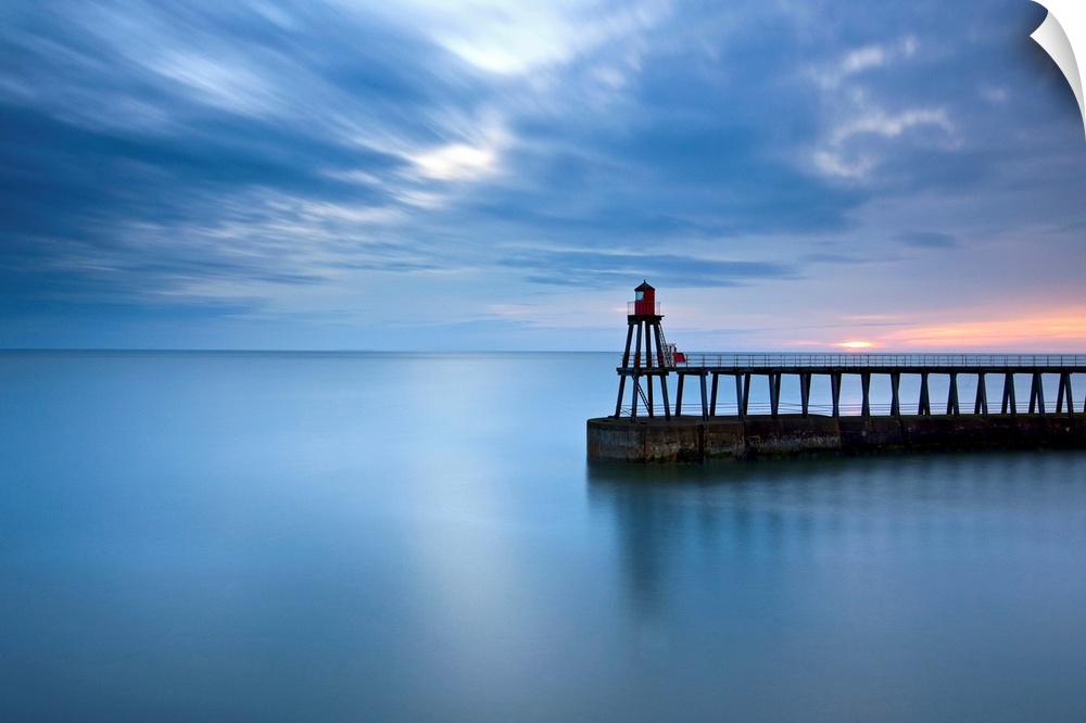 Timeless blue dawn over flat sea with scudding clouds and a lighthouse jetty.